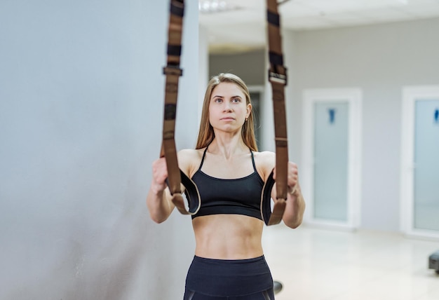 Mulher desportiva fazendo flexões treinando braços com tiras de fitness trx no ginásio em fundo cinza claro Linda mulher musculosa com exercício do sistema trx