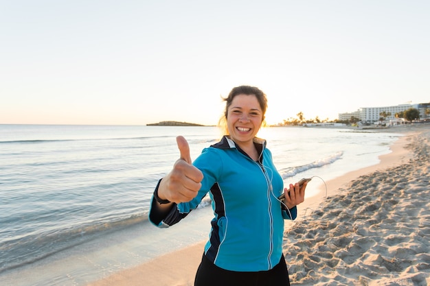 Mulher desportiva motivada fazendo polegares para cima gesto de sucesso após treino urbano na praia.