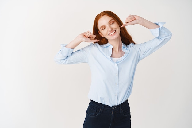 Mulher despreocupada com cabelo ruivo, vestindo blusa, esticando as mãos com sorriso satisfeito e olhos fechados após uma boa soneca, em pé sobre uma parede branca