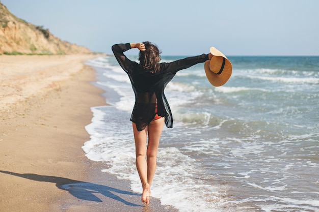 Mulher despreocupada com chapéu andando na praia de areia nas ondas do mar e férias de verão relaxantes