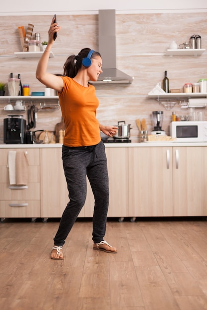 Mulher despreocupada dançando na cozinha ouvindo música em fones de ouvido