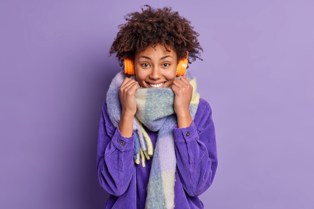 Mulher despreocupada de pele escura com cabelo encaracolado usa lenço quente em volta do pescoço vestidos para clima frio e usa fones de ouvido sem fio para ouvir música enquanto caminha