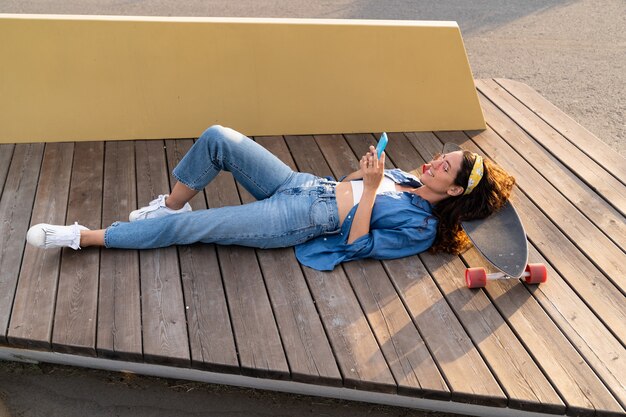 Mulher despreocupada e elegante conversando on-line com o celular e feliz deitada em um longboard ao ar livre