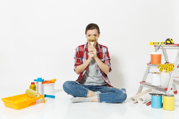 Mulher divertida com roupas casuais, sentada no chão com a escova, instrumentos para quarto de apartamento de renovação isolado no fundo branco. Papel de parede, acessórios para colar, ferramentas de pintura. Conceito de casa de reparo.