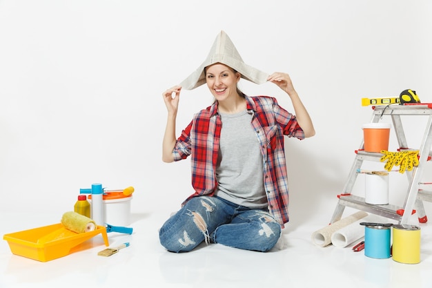 Mulher divertida feliz com chapéu de jornal, sentado no chão com instrumentos para sala de apartamento de renovação isolado no fundo branco. Papel de parede, acessórios para colar, ferramentas de pintura. Conceito de casa de reparo.