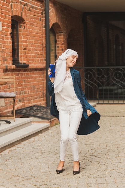 Mulher do Oriente Médio em hijab ouvindo música com fones de ouvido e dançando mulher ao ar livre