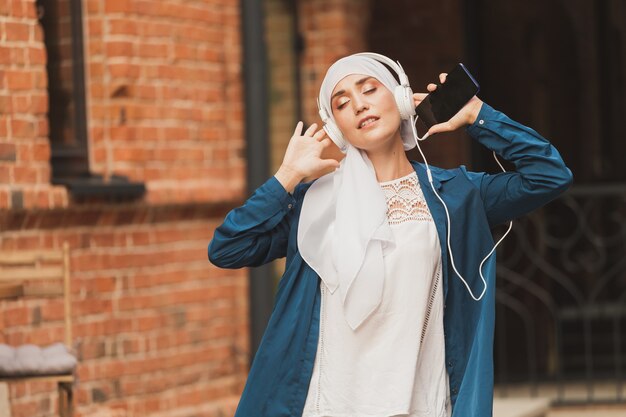 Mulher do Oriente Médio em hijab ouvindo música com fones de ouvido e dançando mulher ao ar livre