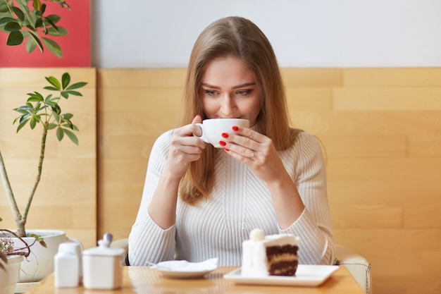 Mulher doce e sincera passa seu tempo no café local, descansa durante o almoço, toma um gole de bebida quente em copo branco.