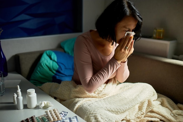 Foto mulher doente com infecções sazonais, alergia a gripe e corrimento nasal, enxugando o nariz, mulher doente coberta com um cobertor, sentada no sofá em casa.