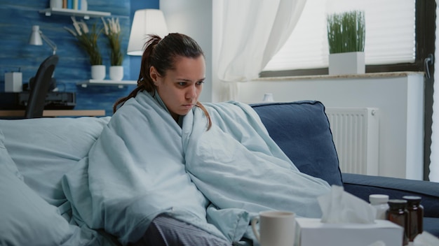 Mulher doente com resfriado usando cobertor para wamth em casa. Pessoa com tremores, febre e calafrios, sentada enrolada em um lençol para curar a infecção do coronavírus com uma xícara de chá e medicamentos.