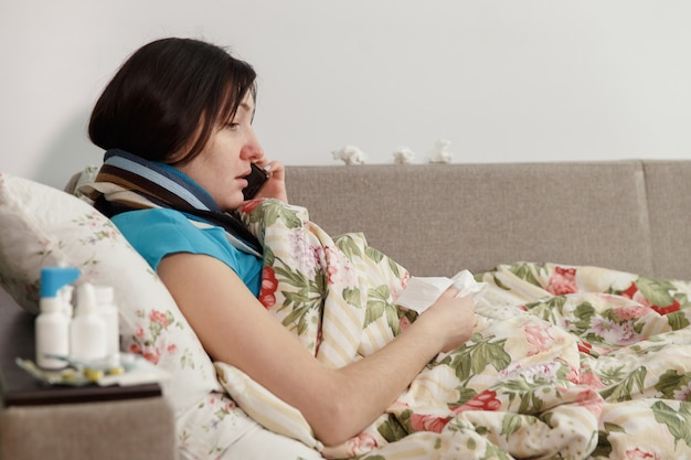 Mulher doente deitada na cama em casa falando ao telefone.