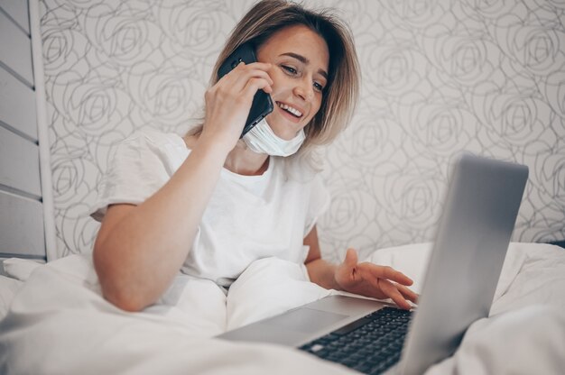 Mulher doente em máscara protetora deitada na cama, falando de smartphone e trabalhando em um laptop durante o isolamento de quarentena em casa Covid-19 vírus pandêmico Corona. Trabalho on-line a distância do conceito de casa.