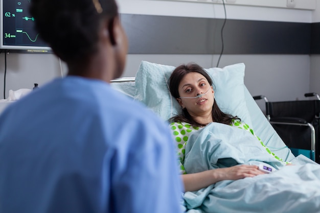 Mulher doente hospitalizada, deitada na cama, enquanto a enfermeira afro-americana verificando o sintoma da doença, escrevendo a perícia médica na área de transferência. Assistente discutindo terapia da doença trabalhando na enfermaria do hospital