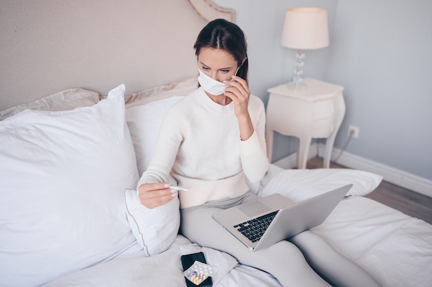 Mulher doente na máscara de proteção facial, deitada na cama com o laptop segurando o termômetro e pílulas em casa