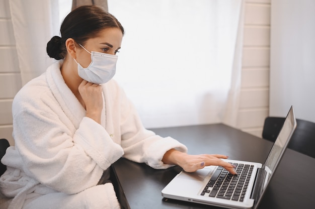 Mulher doente na máscara médica do rosto trabalhando em um laptop durante o vírus de corona pandêmico COVID-19 de isolamento em quarentena em casa. Trabalho on-line a distância do conceito de casa. Sintomas de infecção viral por coronavírus