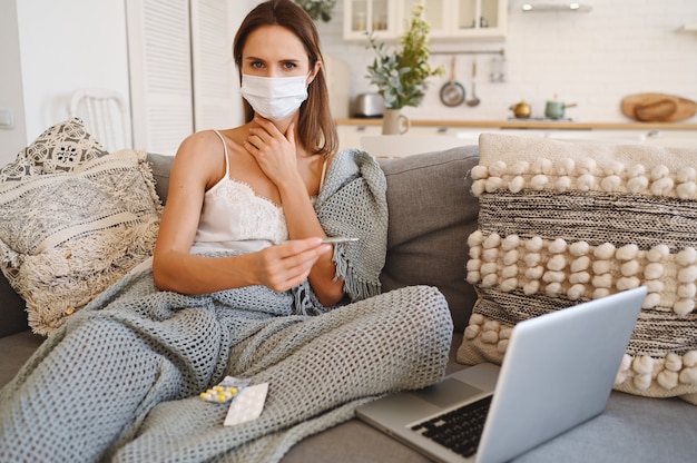 Mulher doente usando máscara de proteção facial, sentado no sofá com o laptop e segurando o termômetro e comprimidos durante o isolamento do auto de quarentena em casa.