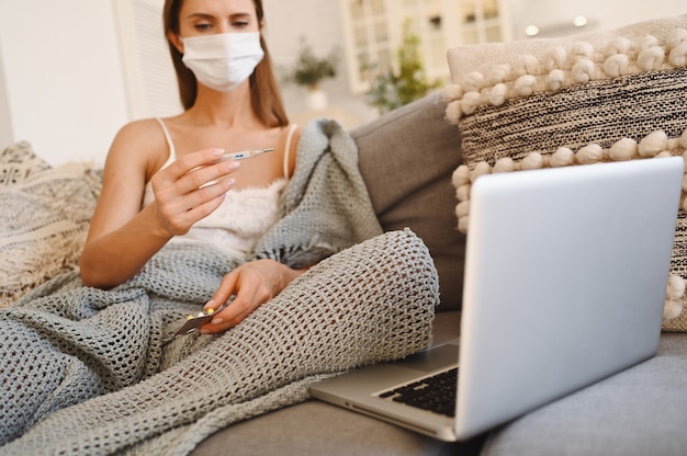 Mulher doente usando máscara de proteção facial, sentado no sofá com o laptop e segurando o termômetro e comprimidos durante o isolamento do auto de quarentena em casa.