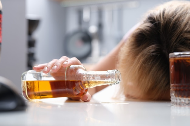 Foto mulher dormindo à mesa com uma garrafa de álcool nas mãos, closeup