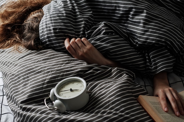 Mulher dormindo coberta sob o cobertor na cama e vintage despertador no travesseiro. manhã preguiçosa ana acordar conceito