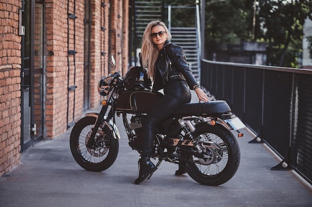 Mulher dramática sexy está estacionando sua moto nova brilhante ao lado da parede de tijolos.
