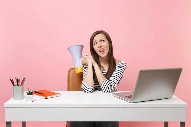 Mulher duvidosa perplexa olhando para cima mostrando a língua segurando o megafone sentar no trabalho na mesa branca no escritório com laptop pc isolado em fundo rosa pastel. Conceito de carreira empresarial de realização. Copie o espaço.