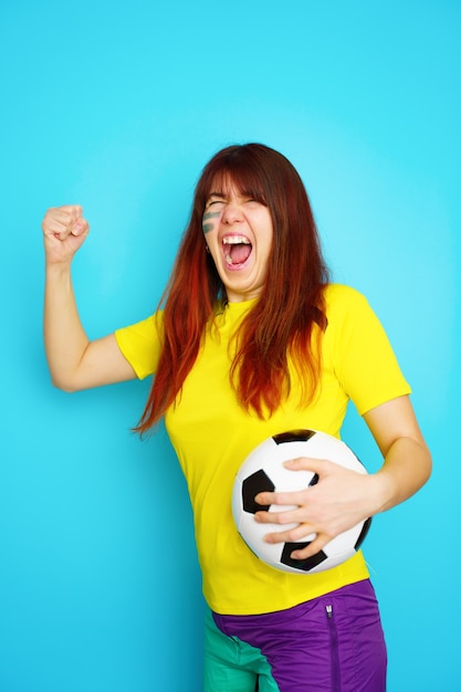 Mulher é fã de futebol com uma camiseta amarela e uma bola de futebol no fundo azul