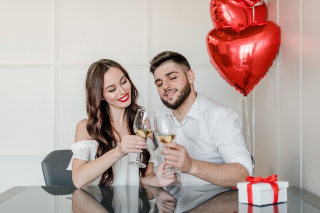 Mulher e homem bonito feliz bebem vinho branco em casa com presente na caixa de presente e balões em forma de coração vermelho em casa