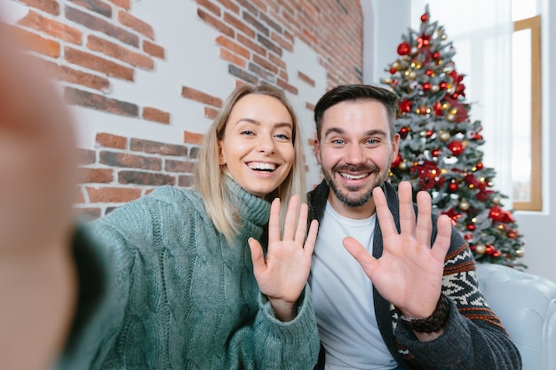 Mulher e homem em casa olhando para a câmera do smartphone cumprimentando amigos com feriados, uma jovem família durante as férias de ano novo e natal