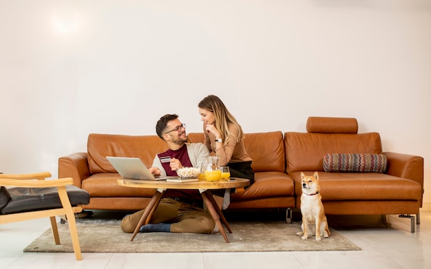 Mulher e homem jovem usando laptop para pagamento on-line enquanto estão sentados no sofá com seu cachorro shiba inu em casa
