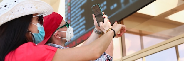 Mulher e homem usando máscaras de proteção médica tiram fotos de um painel de informações na estação de trem