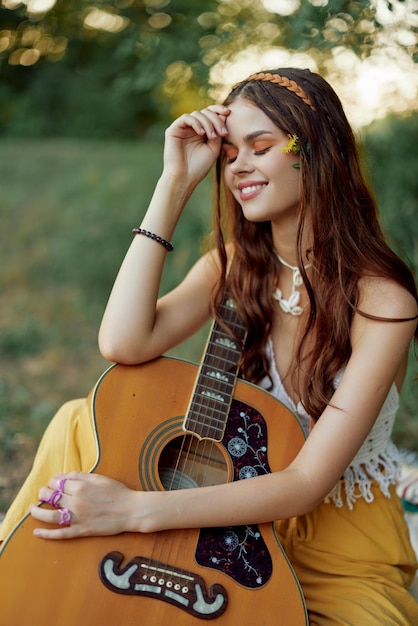 Mulher ecológica hippie com uma guitarra relaxando na natureza sentada em uma manta à beira do lago à noite sob os raios do sol poente Um estilo de vida em harmonia com o corpo e a natureza