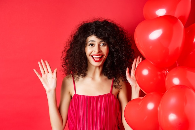 Mulher elegante animada com cabelo encaracolado, usando vestido, levantando as mãos e rindo feliz, em pé perto de balões de coração, fundo branco.