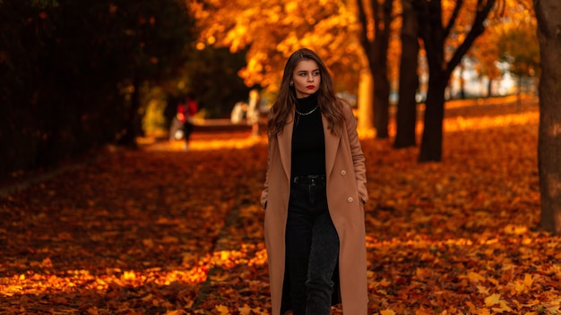 Mulher elegante com lábios vermelhos em um casaco bege elegante com um suéter preto caminha em um parque de outono com folhagem laranja ao pôr do sol