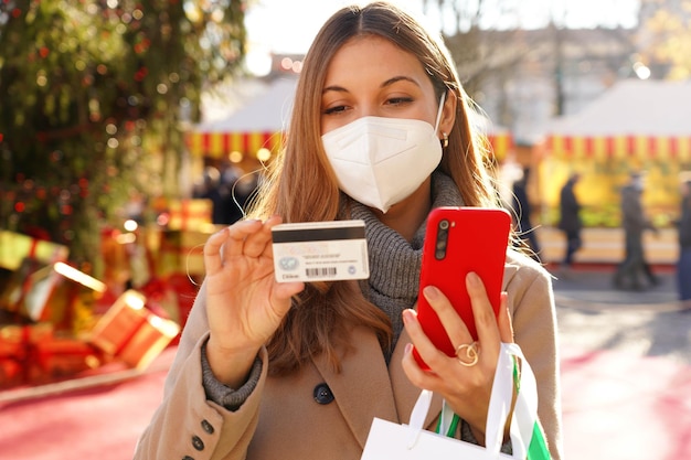 Mulher elegante com máscara protetora KN95 FFP2 observando o número do cartão de crédito para inserir no smartphone para pagamento online com mercados de Natal no fundo