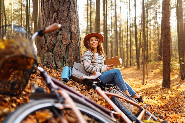 Mulher elegante com um livro perto da árvore gosta do clima de outono no parque ao fundo da bicicleta