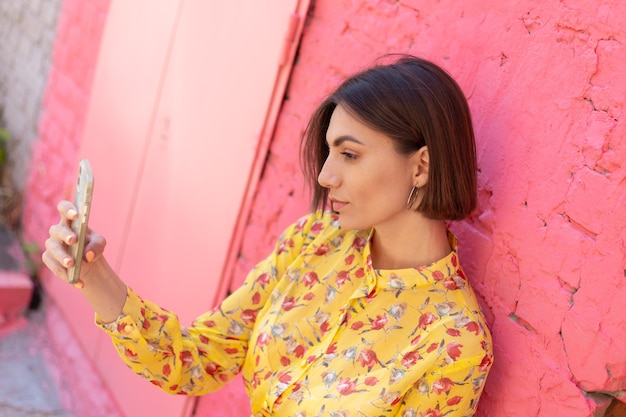 Mulher elegante com vestido amarelo de verão na parede de tijolo rosa feliz, positivo, tirar selfie no celular