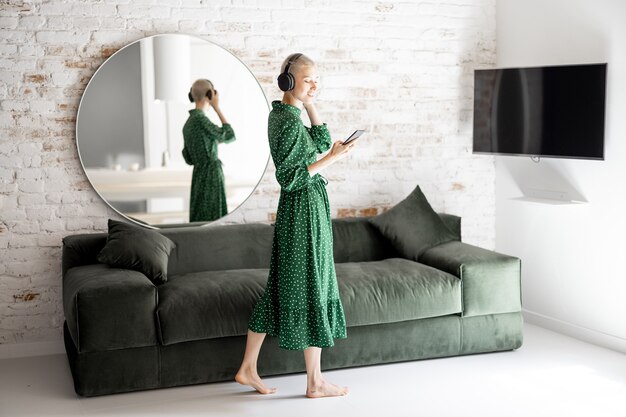 Mulher elegante com vestido verde aprecia a música com fones de ouvido e celular dançando na sala de estar em casa
