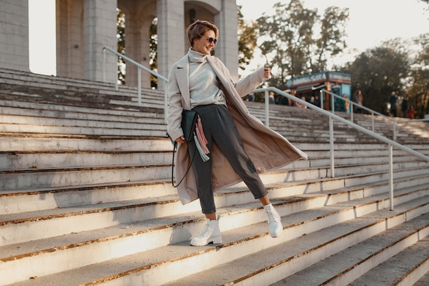 Mulher elegante, confiante e elegante andando na rua com um casaco de estilo elegante, usando óculos escuros, bolsa e botas brancas