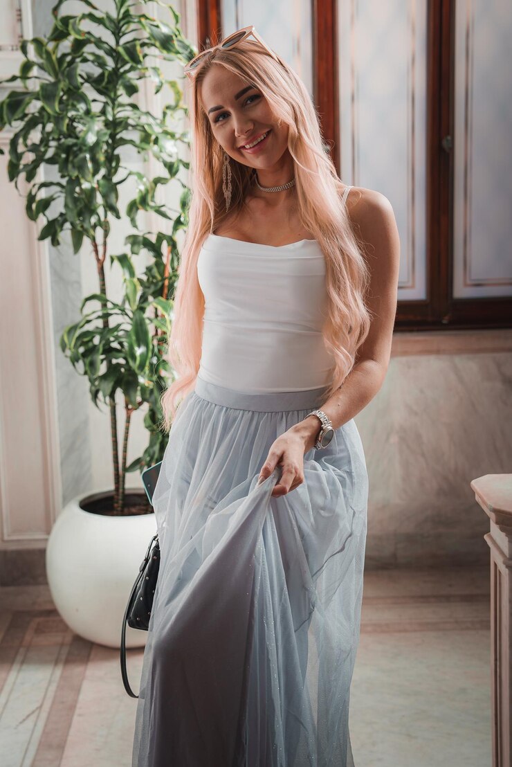 Foto mulher elegante de blusa branca e saia de tulha cinza posando em uma sala bem iluminada com planta em vaso