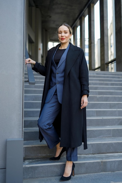 Mulher elegante de terno e casaco posando nas escadas de um prédio