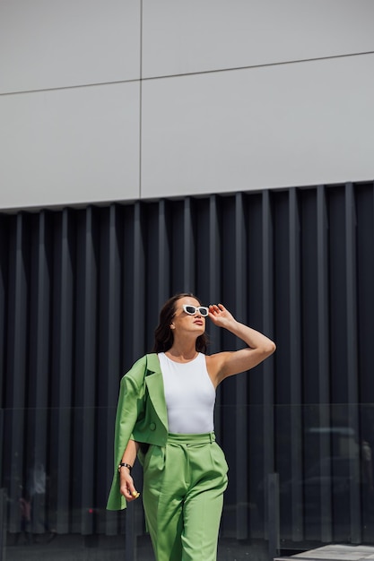 Mulher elegante e confiante de moda usando óculos de sol rosa de moda, lenço de seda, blazer com saco verde, posando na rua de uma cidade europeia, retrato de moda ao ar livre, copie o espaço vazio para o texto