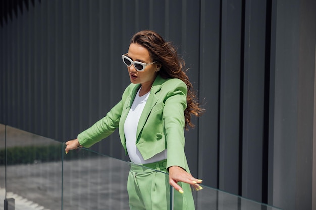 Mulher elegante e confiante de moda usando óculos de sol rosa de moda, lenço de seda, blazer com saco verde, posando na rua de uma cidade europeia, retrato de moda ao ar livre, copie o espaço vazio para o texto