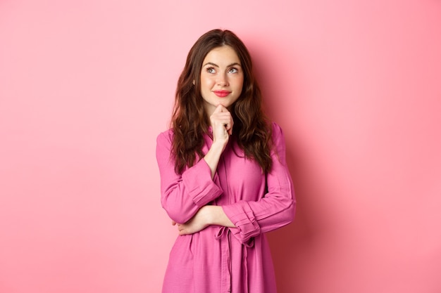 Mulher elegante e pensativa com maquiagem e vestido de primavera, olhando de lado para o logotipo e sorrindo satisfeito, pensando em pé contra a parede rosa.