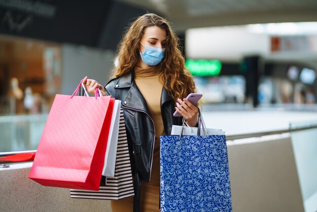 Mulher elegante em máscara protetora médica com sacolas de compras, usando seu telefone.