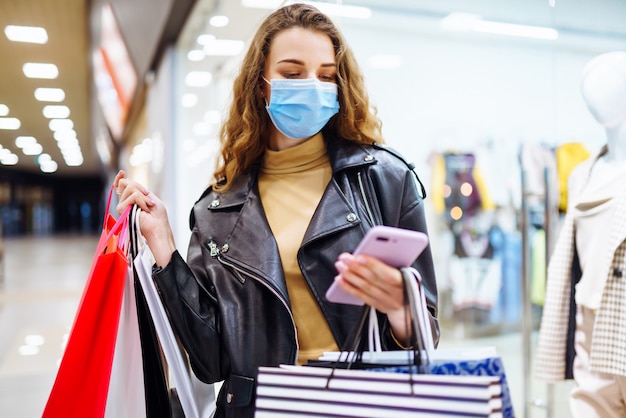 Mulher elegante em máscara protetora médica com sacolas de compras, usando seu telefone.