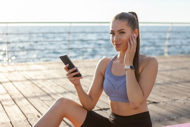 Mulher elegante em roupas esportivas usa um smartphone e ouve música com fones de ouvido enquanto está sentada em um tapete na praia ao nascer do sol