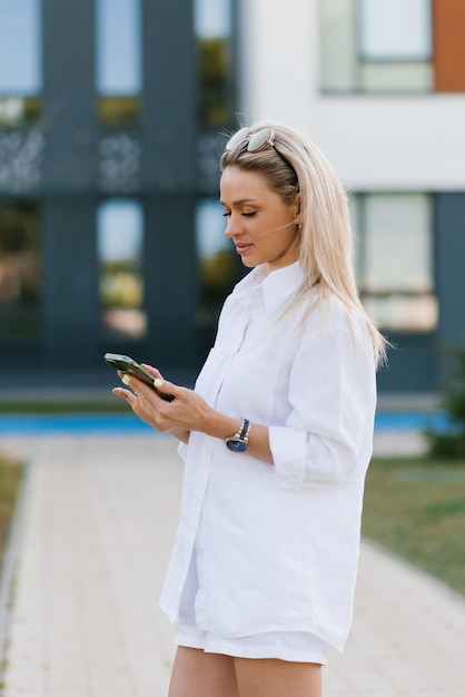 Mulher elegante em um terno de linho branco com shorts e uma camisa está comprando online em um smartphone enquanto caminhava pela cidade no verão