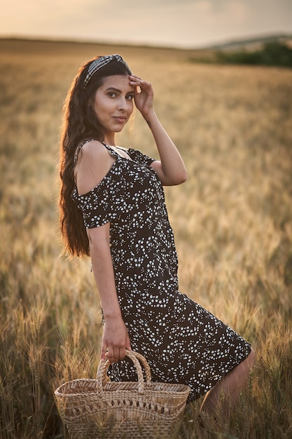 Mulher elegante em um vestido com cesta no campo de trigo ao pôr do sol.