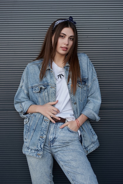 Foto mulher elegante em uma jaqueta jeans e jeans posando contra uma parede cinza na rua.