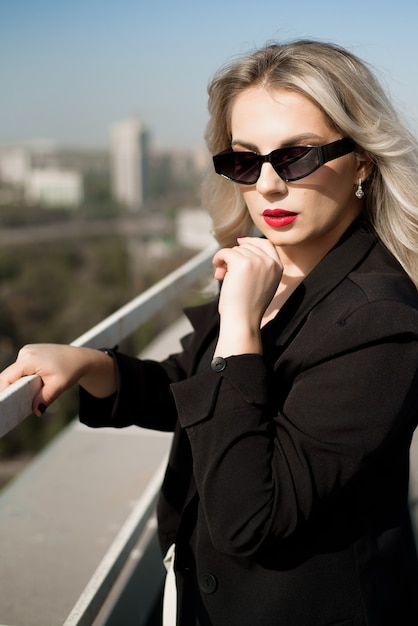 Mulher elegante em uma jaqueta preta e óculos escuros no terraço do bar da cobertura, olhando para a cidade.